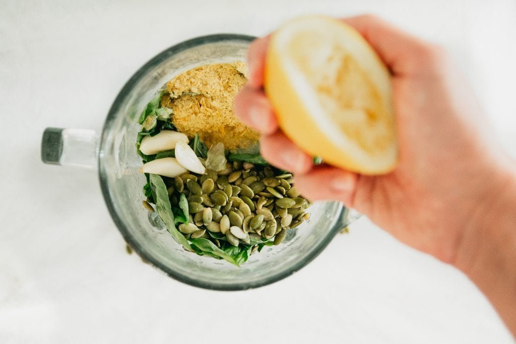 A hand squeezes juice from a lemon into a blender which contains other ingredients for a vegan pesto recipe.