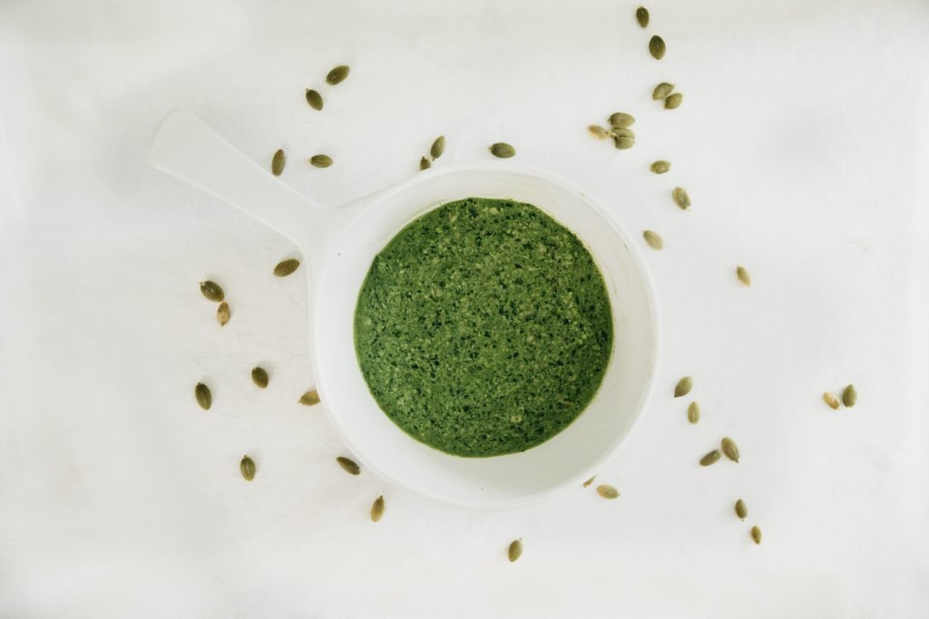 A white bowl with basil pesto sauce in it surrounded by various seeds